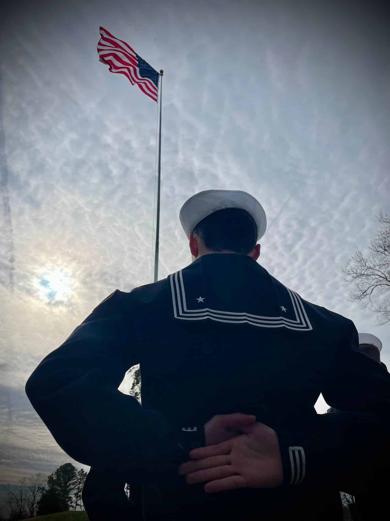 Cadet in dress blues facing an American flag against a dramatic sky