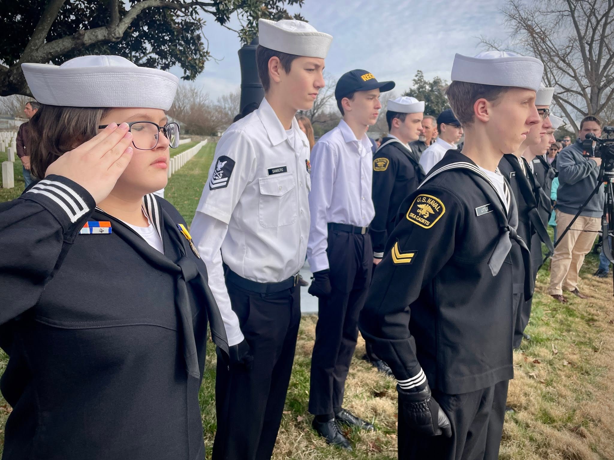 Female cadet saluting at a cemetery ceremony as media cameras roll